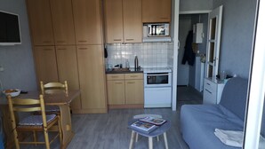 A compact kitchen with wooden cabinets, a microwave, and a built-in oven. There's a dining table with chairs and a small table with a magazine. A sofa is in the living area.