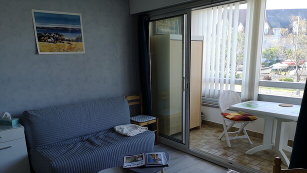 A modern living room with a blue sofa, a small table, and a chair. There is a sliding glass door leading to an outdoor balcony with a white table and chairs.
