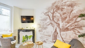 Un salon avec une cheminée, un canapé agrémenté d’un coussin jaune, un fauteuil en osier et une fresque murale représentant un arbre.