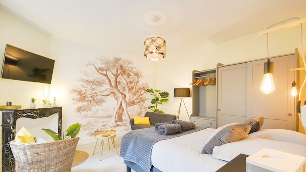 Une chambre moderne avec un grand lit, un téléviseur à écran plat, un téléviseur fixé au mur, un panier en osier, une plante, un bureau avec une lampe et une armoire.