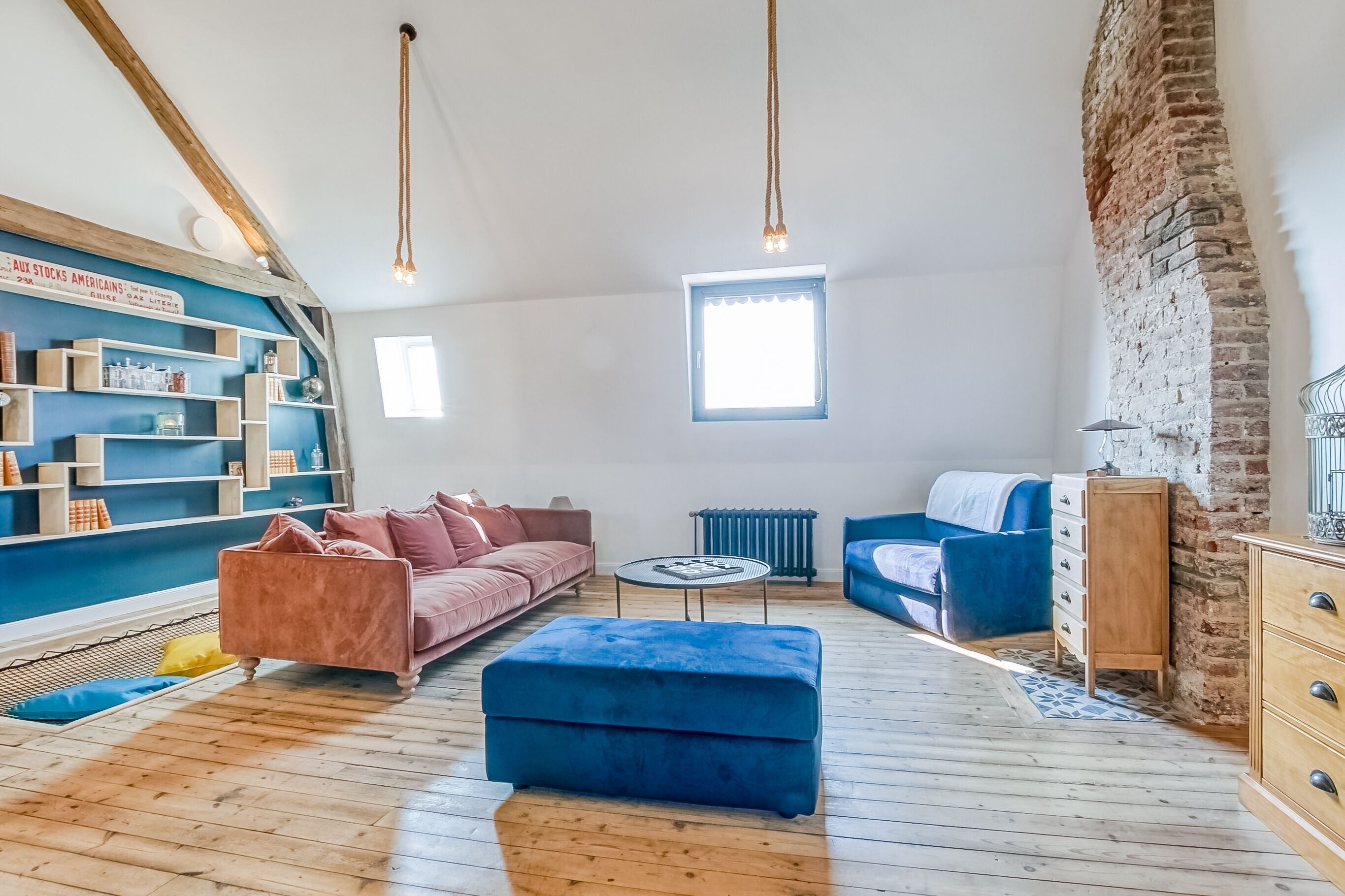 Een kamer met een loft-plafond, een blauwe bank, een roze bank, een blauwe poef, een klein rond tafeltje en een bakstenen muur.