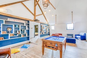 Une pièce spacieuse équipée d’une table de billard, de canapés bleus, de bibliothèques en bois et d’un plafond haut avec des poutres apparentes.