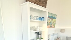 Ein weißes Schränkchen mit Regalen, ein kleiner weißer Tisch, eine Lampe und ein Bild an der Wand.