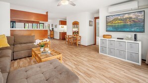 A living room with a sofa, coffee table, and TV. There is a dining area with chairs and a table. The room has wooden flooring and a ceiling fan.