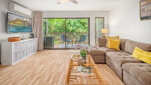 A modern living room with a sofa, a coffee table, a TV, and a balcony with a view of greenery.