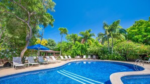 A swimming pool with lounge chairs and umbrellas, surrounded by lush greenery and palm trees.