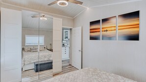 Une chambre à coucher avec un lit, un miroir, un ventilateur de plafond et un mur orné d’un tableau représentant un coucher de soleil.