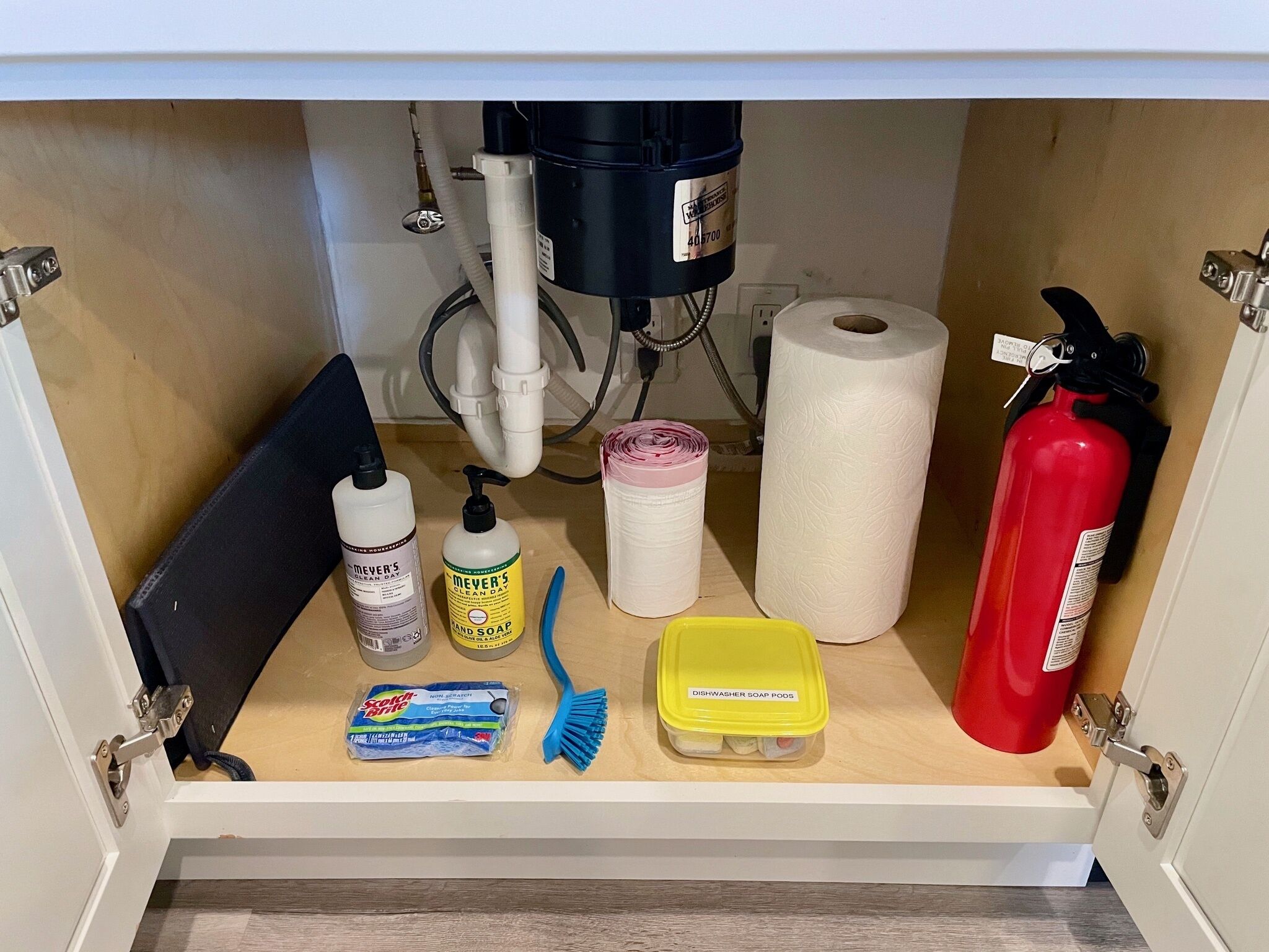 Ein Küchenschrank mit verschiedenen Reinigungsmitteln, darunter Geschirrspülmittel, Handseife, eine Zahnbürste, ein Behälter für Spülmittel, eine Küchenrolle und ein Feuerlöscher.