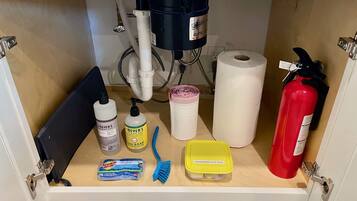 Ein Küchenschrank mit verschiedenen Reinigungsmitteln, darunter Geschirrspülmittel, Handseife, eine Zahnbürste, ein Behälter für Spülmittel, eine Küchenrolle und ein Feuerlöscher.