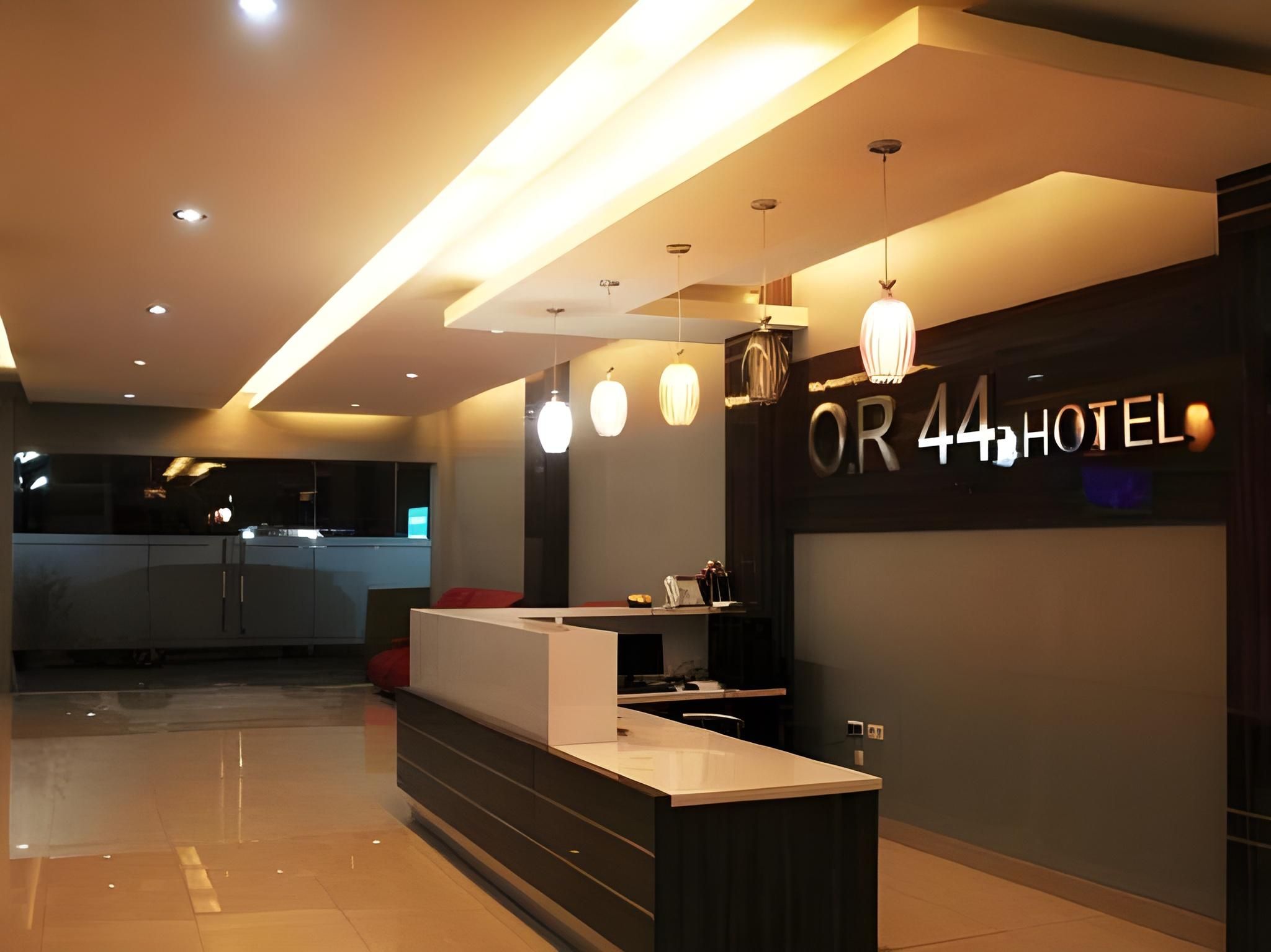 A modern hotel reception area with a counter, pendant lighting, and a visible sign for 'OR 44 HOTEL'.