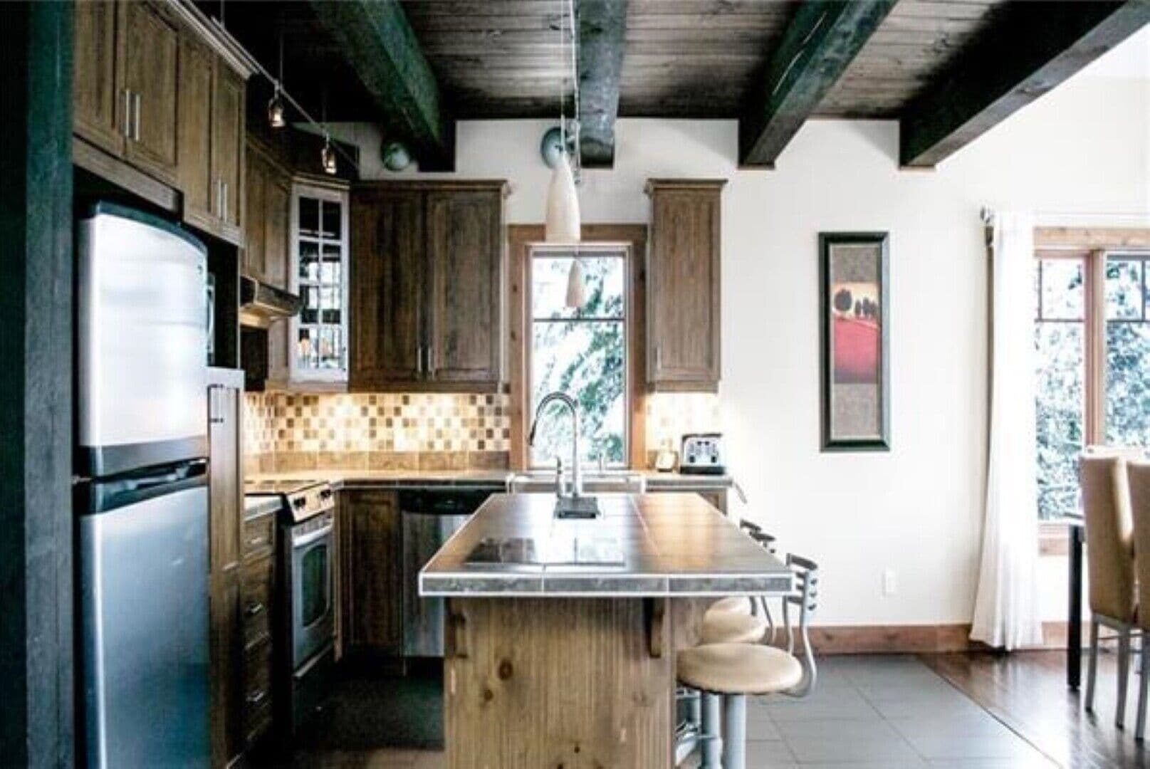 Une cuisine dotée de meubles en bois, d’un réfrigérateur en acier inoxydable, d’un pare-feu en carrelage et d’un coin bar avec des tabourets.