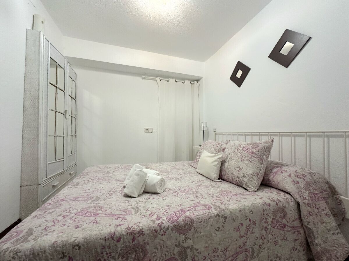 Una cama bien hecha con una colcha y cojines estampados con motivos florales, un armario blanco y un radiador blanco en la pared.