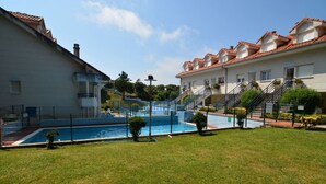 A swimming pool surrounded by a fence, with a building featuring a red-tiled roof in the background. - Apartamento con piscina junto a la playa - La Aren (Arnuero)