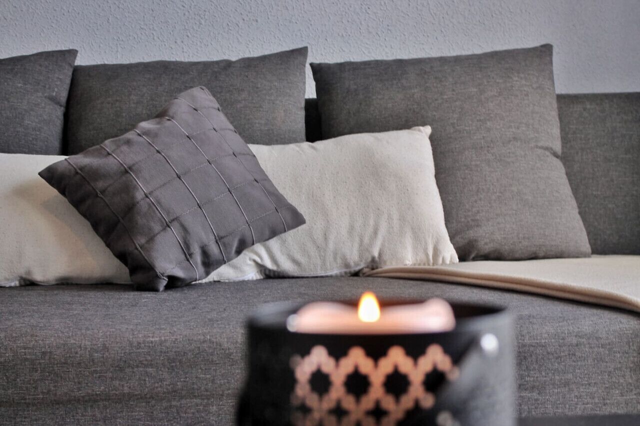 Ein graues Sofa mit einem schwarz-weißen Kissen, eine Kerze in einem schwarzen Kerzenhalter und eine graue Wand im Hintergrund.