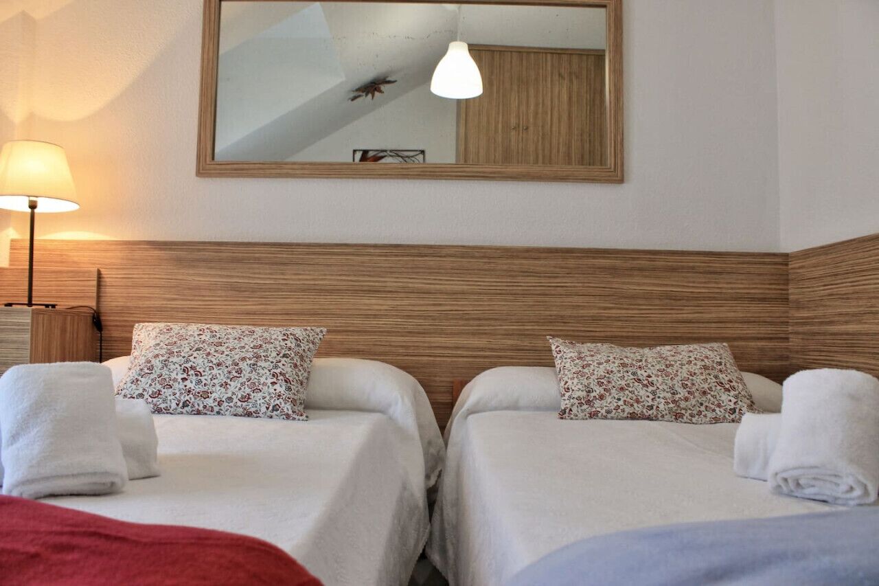 Une chambre d’hôtel avec deux lits, chacun agrémenté d’un coussin à motifs floraux et d’une serviette blanche. On y trouve une tête de lit en bois, un miroir au mur et une lampe sur le côté gauche.