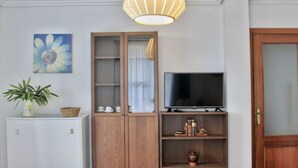 A room with a television on a wooden stand, a cabinet with glass doors, a potted plant, and a painting of a flower on the wall.