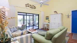 A living room with a sofa, armchair, and a dining table. There is a ceiling fan, a cabinet, and a blue door. - A Peace of Heaven (Sanibel)
