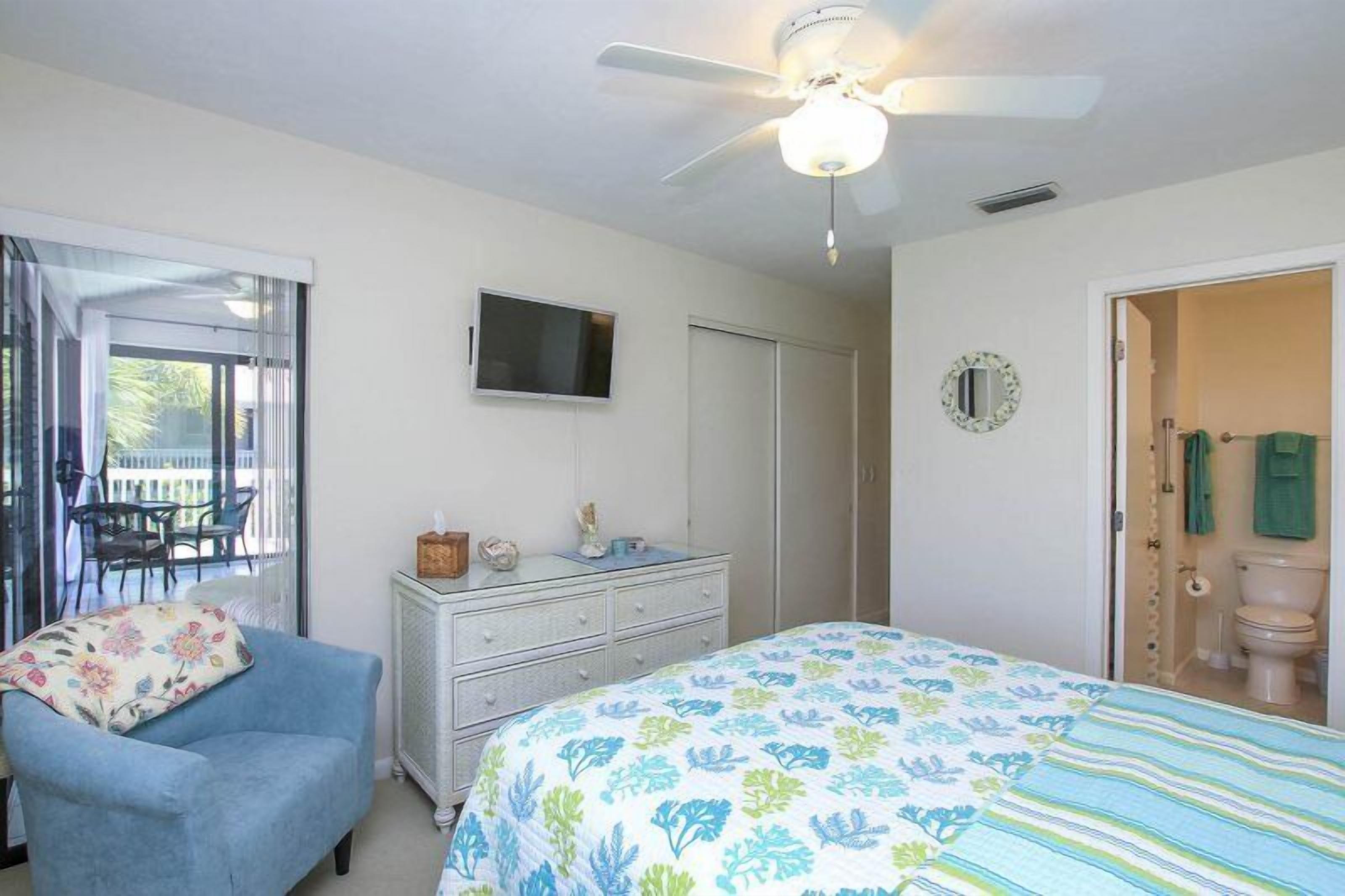 A bedroom with a bed, a dresser, a television, a ceiling fan, a window, and a door leading to a bathroom.