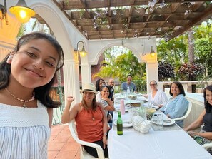 Eine Gruppe von Menschen speist an einem Tisch im Freien, überdacht von einer Pergola, umgeben von üppigem Grün.
