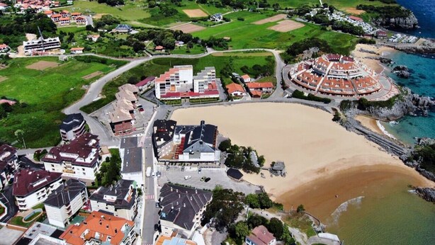 Vista aérea de un pueblo costero con una playa central, edificios residenciales circundantes y una estructura circular con tejados rojos cerca del agua.