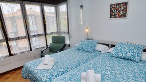 Habitación de hotel con dos camas, una silla, un ventanal con cortinas de encaje, un cuadro en la pared y una mesita de noche con lámpara.