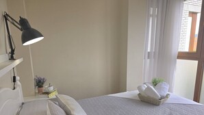 A neatly made bed with pillows, a bedside lamp, a small table with a plant, and a basket of towels.