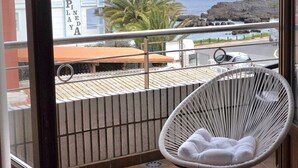 A balcony with a wicker chair, a view of the ocean, and a building with a sign reading 'HOTEL PINE LA DYA'.