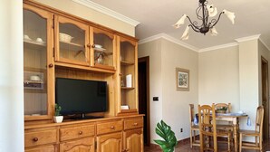 A living room with a wooden cabinet, a television, a dining table with chairs, and a chandelier. - Apartamento con vistas al mar - Pineda 6404 (Noja)