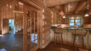 A log cabin interior with a wooden staircase, a kitchen area with a bar, and a large window.
