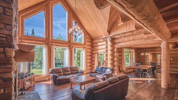 A spacious log cabin interior with large windows, a fireplace, leather sofas, and a dining area.