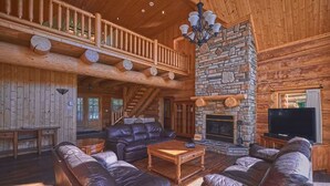 A log cabin interior with a stone fireplace, leather sofas, a wooden dining table, and a TV.