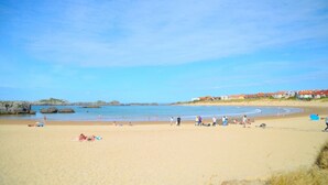 A wide sandy beach with people sunbathing and swimming, a clear blue sky, and buildings in the background.