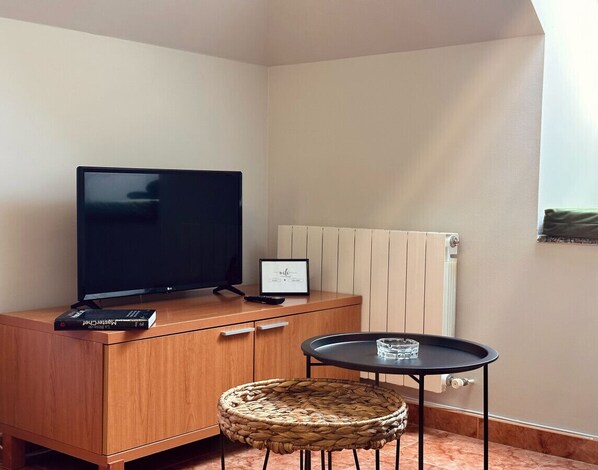 Een kamer met een hoekopstelling, voorzien van een houten kast, een flatscreen televisie, een klein rond tafeltje, een rieten stoel en een lijst met een afbeelding aan de muur.