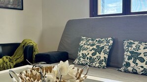 Ein Wohnzimmer mit grauem Sofa, zwei gemusterten Kissen, einer grünen Decke und einer dekorativen Schale auf einem Glastisch.