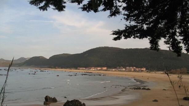 Ein Strand mit sandigem Ufer, vereinzelten Felsen und einer entfernten Hügelkette mit Gebäuden.