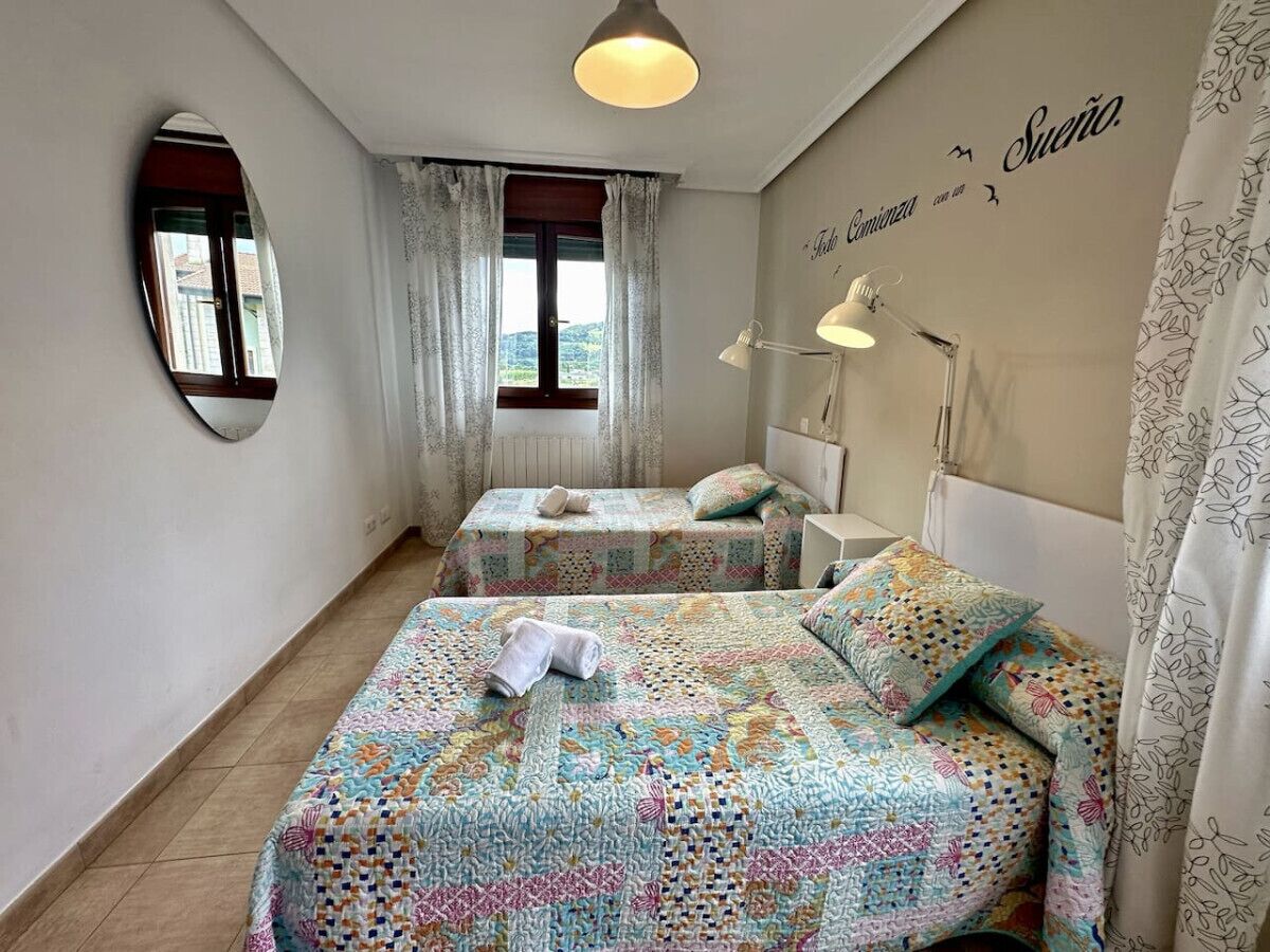 A bedroom with two beds, a window with curtains, a wall clock, and a ceiling light.