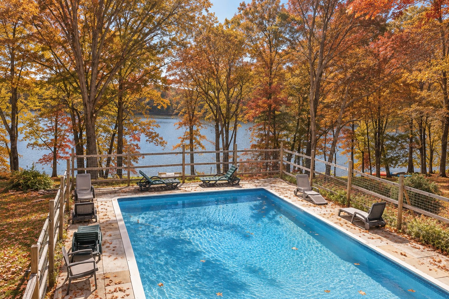 A rectangular swimming pool with a wooden deck and lounge chairs, surrounded by trees with autumn foliage, and a view of a body of water.