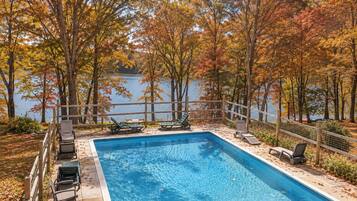 A rectangular swimming pool with a wooden deck and lounge chairs, surrounded by trees with autumn foliage, and a view of a body of water.
