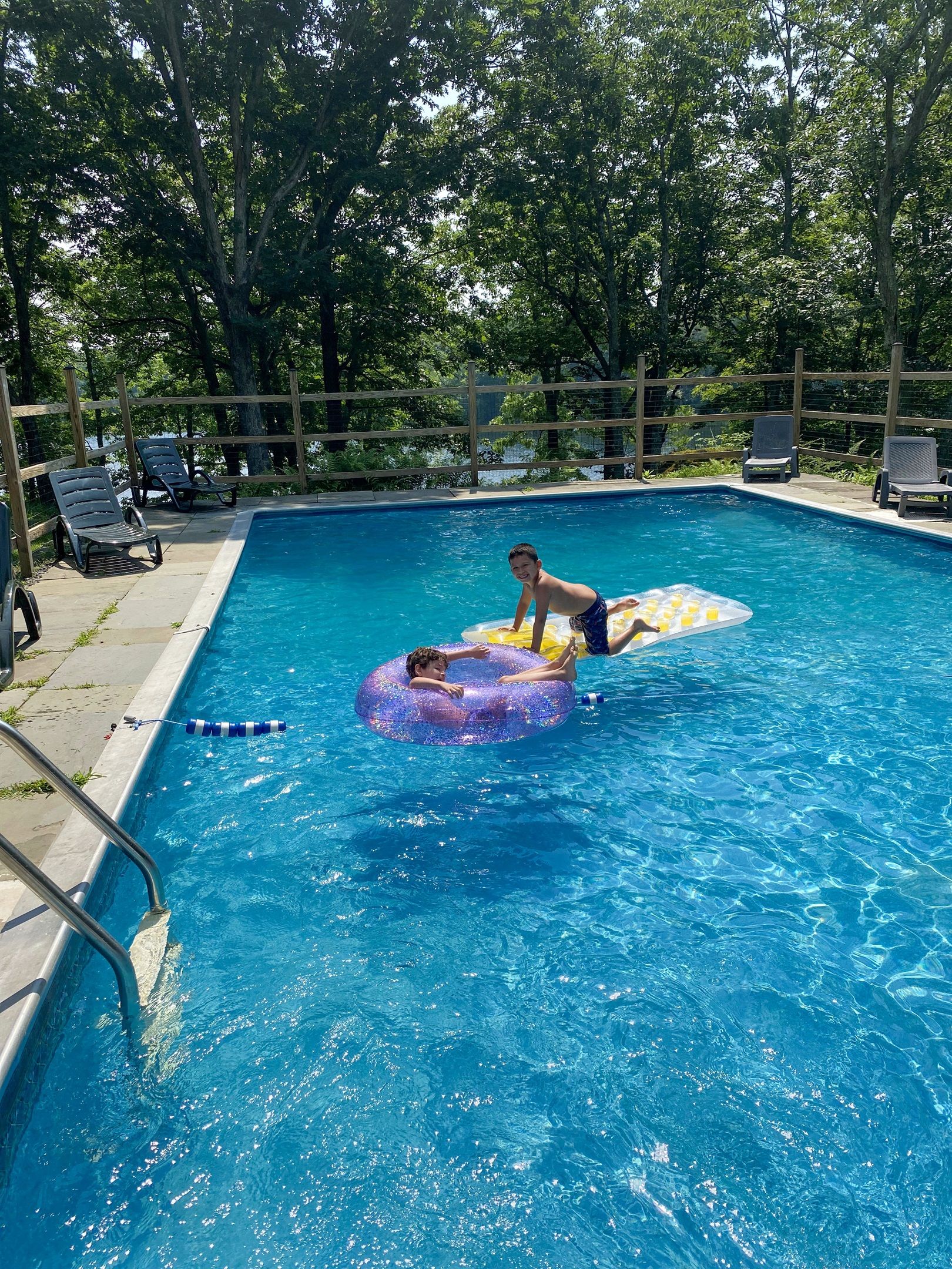 Une piscine avec un enfant sur un flotteur bleu, entourée de chaises longues et d’une clôture en bois.
