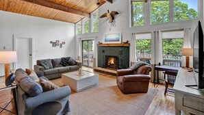 A spacious living room with a fireplace, large windows, and a high ceiling with exposed beams. - Lakefront Retreat, Private Pool, Hot Tub, Game Rm, Ping Pong, Foosball (Bushkill)