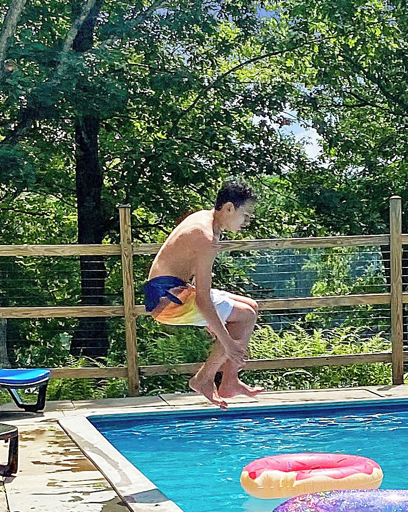 Eine Person springt in einen Swimmingpool, im Hintergrund ein Holenzaun und Grünpflanzen.