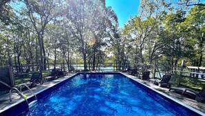 Une piscine entourée d’arbres, avec des chaises longues et une terrasse en bois, sous un ciel dégagé.