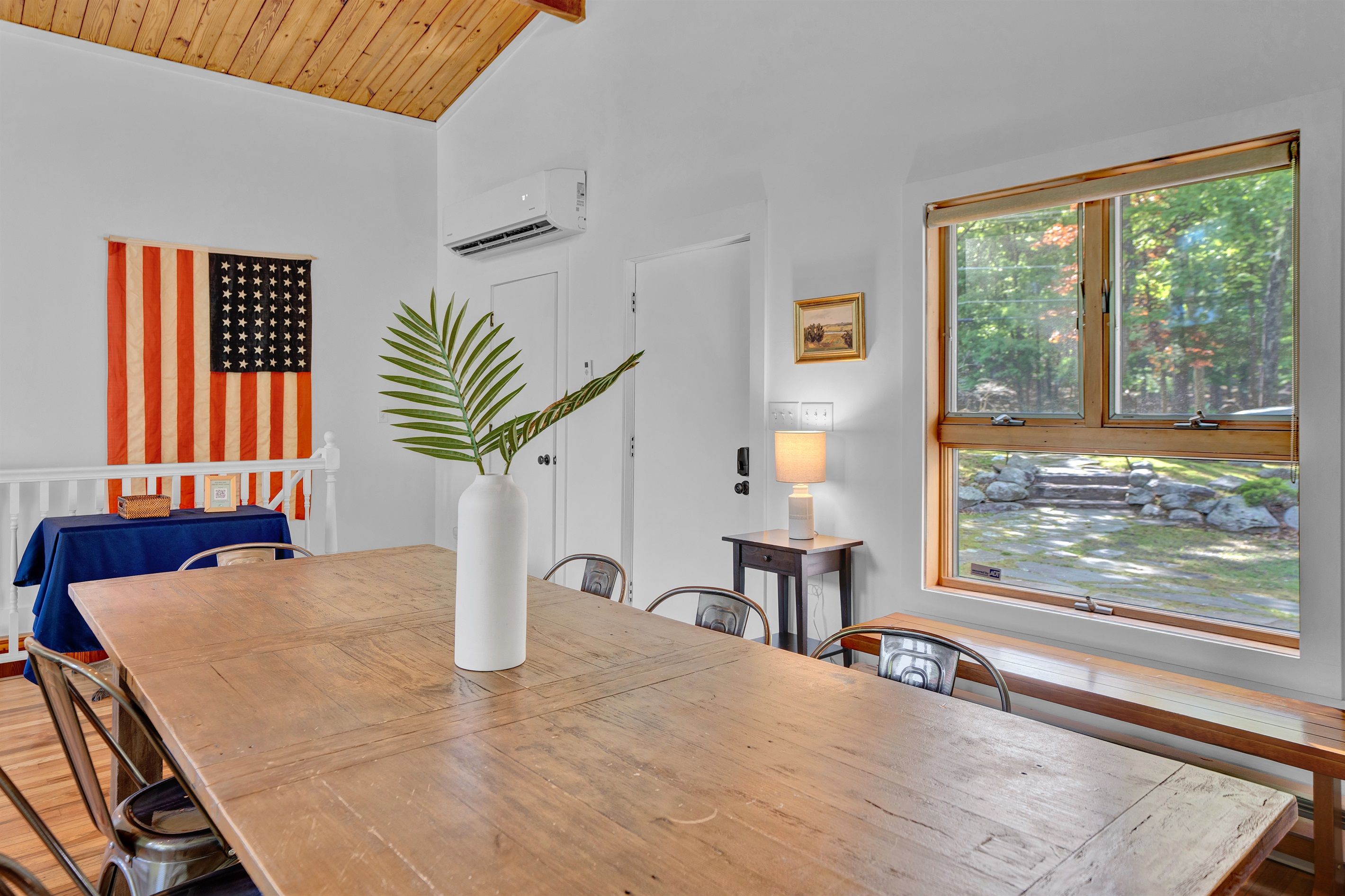 Ein Essbereich mit Holztisch, Stühlen, einer Vase mit Pflanze, einer amerikanischen Flagge und einem Fenster mit Blick auf Bäume.
