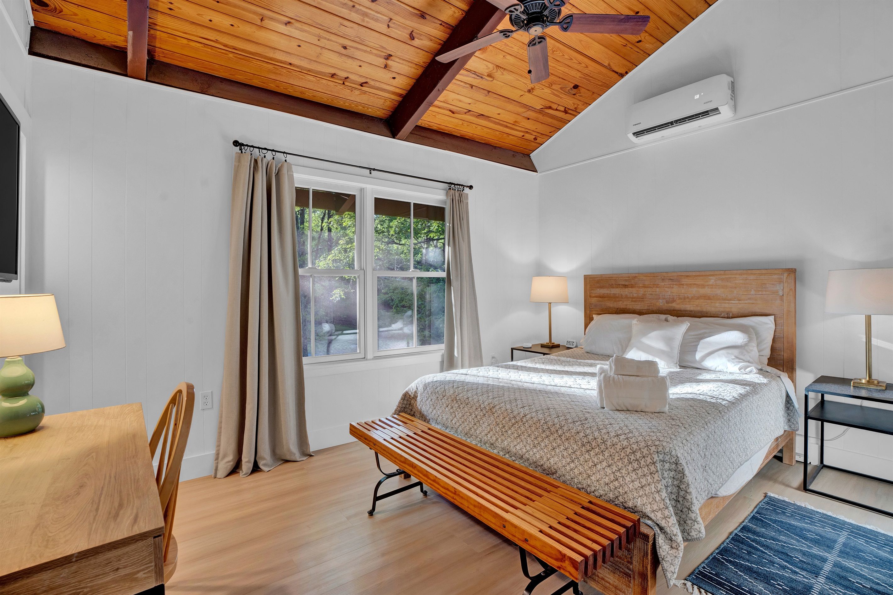 A bedroom with a bed, bedside lamps, a wooden bench, a desk, and a window with curtains.