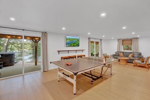 A spacious room with a ping pong table, a sofa, and a large window overlooking a forested area. - Lakefront, Hot Tub, Beach, Free Boats, Pets OK, Fully Loaded, Dock (Bushkill)