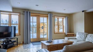 A modern living room with a flat-screen TV, leather sofa, wooden coffee table, and large windows with curtains.