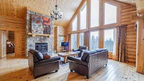 A log cabin interior with a stone fireplace, two sofas, a coffee table, and large windows offering a view of the outdoors.