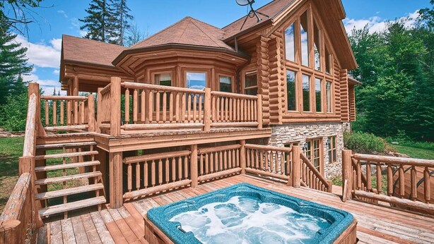 A log cabin with a hot tub on the deck, a satellite dish, and a stone foundation.