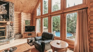 A log cabin interior with a stone fireplace, a television, a leather chair, a round wooden table, and large windows overlooking a swimming pool.