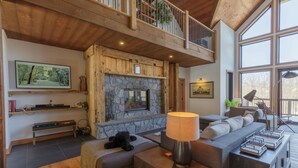 A spacious living room with a stone fireplace, a large window, and a high ceiling with wooden beams. - Laurentian Chalet: 4K Cinema, Sauna & Hot Tub (Morin-Heights)